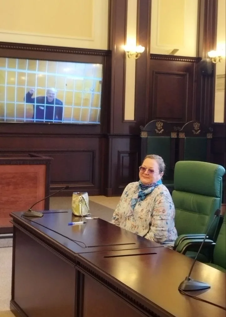 Александр Скобов на экране видеосвязи за решёткой; женщина сидит за столом в зале суда.